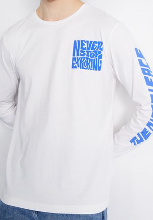 Langärmeliges weißes T-Shirt mit blauem Schriftzug "NEVER STOP EXPLORING" auf der Brusttasche und "THE NORTH FACE" entlang des Ärmels.