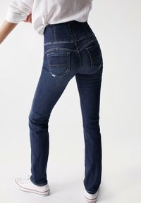 Jeans de mezclilla oscuros de tiro alto con una textura suave, diseño de cinco bolsillos y detalles de costura visibles, combinados con zapatillas blancas.