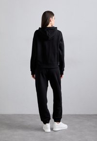 Crna kapuljača i odgovarajući trenirka izrađeni od mekog materijala, sa rebrastim manžetama i opuštenim krojem. Bijele tenisice upotpunjuju look.