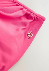 Rozā audums ar gludu tekstūru, kas izceļas ar savāktu dizainu un nelielu zelta logotipu sānos.