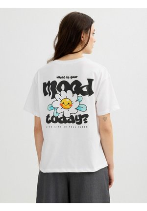 Mujer con cabello largo y castaño que lleva una camiseta blanca con una flor sonriente y el texto "¿Cuál es tu estado de ánimo hoy? Vive la vida en plena floración."