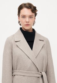 Ungdom med kort, krøllete hår iført beige belted frakk over svart ribbestrikket turtleneck-genser, ansiktet vendt fremover mot hvit bakgrunn.