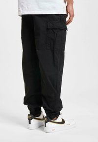 Schwarze Cargo-Hosen mit Seitentaschen, elastischen Bündchen und lässigem Schnitt, kombiniert mit weißen Sneakers mit schwarz-goldenen Akzenten.