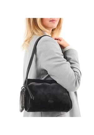 Frau in einem grauen Blazer, die eine schwarze, flauschige Umhängetasche mit Reißverschluss-Details über der Schulter trägt, vor weißem Hintergrund.