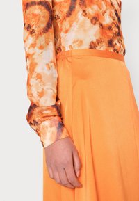 Langärmliges Hemd mit Batikmuster in Orange und Braun, kombiniert mit einem einfarbigen orangen, plissierten Rock, der glatte Texturen präsentiert.