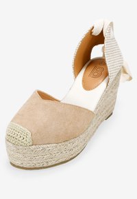 Cuña de esparto beige con parte superior de suave gamuza, suela de yute trenzado y correa de talón de tela texturizada con un lazo decorativo.