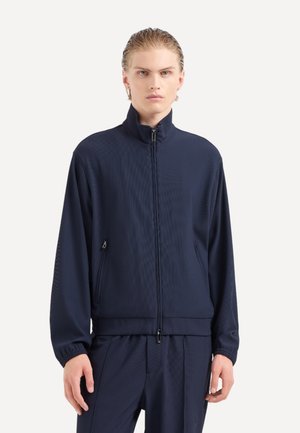 Giacca sportiva blu navy con alto collo, chiusura zip, tessuto strutturato e tasche laterali. Dotata di polsini elastici e design aderente.