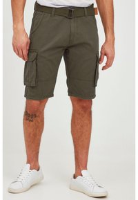 Man wearing olive green cargo shorts, white sneakers, and a canvas belt, standing with tattoo visible on left lower leg.