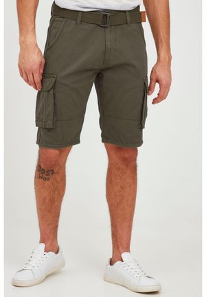 Man som bär olivgröna cargoshorts, vita sneakers och ett canvasbälte, står med en tatuering synlig på vänster underben.