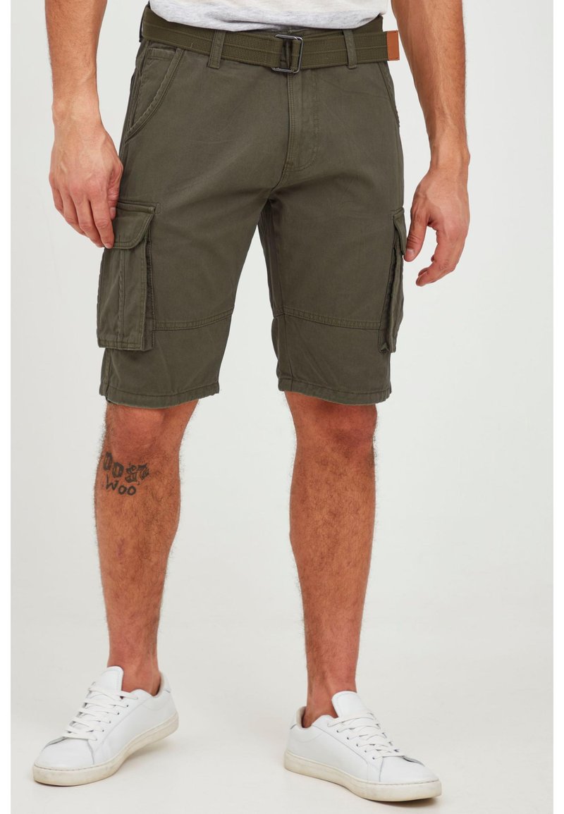 Man wearing olive green cargo shorts, white sneakers, and a canvas belt, standing with tattoo visible on left lower leg.