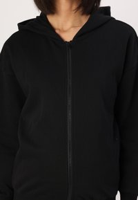 Felpa nera con zip, realizzata in tessuto testurizzato, con zip frontale e cappuccio regolabile con cordino. Nessun motivo o dettaglio visibile.