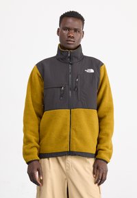 Giacca in pile nera e gialla con colletto alto, chiusura con zip, due tasche sul petto e logo The North Face sul lato sinistro.