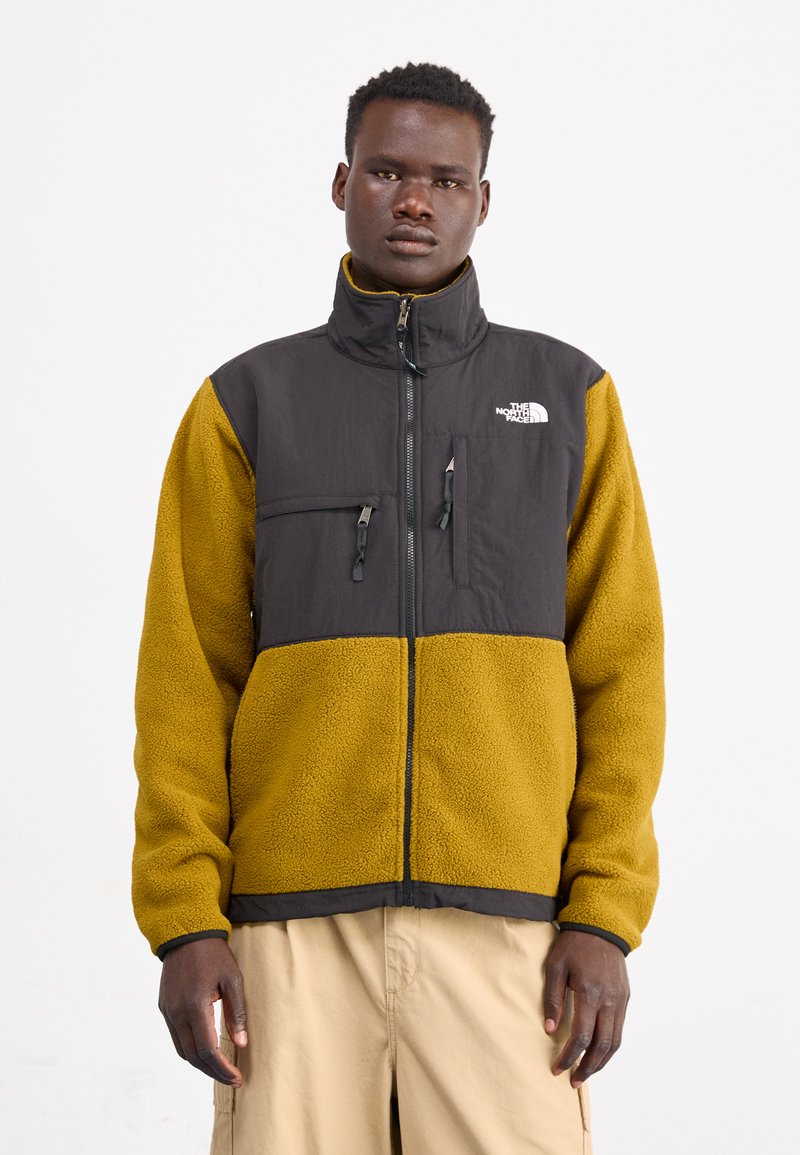 Giacca in pile nera e gialla con colletto alto, chiusura con zip, due tasche sul petto e logo The North Face sul lato sinistro.