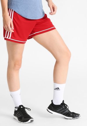 Osoba w czerwonych sportowych szortach, niebieskiej koszulce, białych skarpetkach Adidas i czarnych butach sportowych Adidas, stojąca na białym tle.