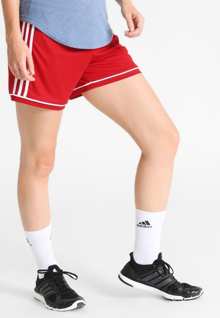 Personne portant un short de sport rouge, un t-shirt bleu, des chaussettes blanches Adidas et des baskets noires Adidas, debout sur un fond blanc.