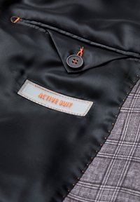 Schwarzer Satin-Stoff mit einem orangefarbenen Knopf und einem Etikett mit der Aufschrift "ACTIVE SUIT". Grauer Karostoff ist am Rand sichtbar.