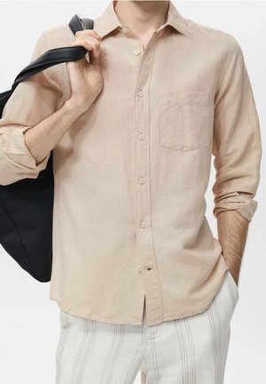 Mand iført en beige skjorte med knapper og opkørte ærmer og hvide stribede bukser, med en sort taske over skulderen.