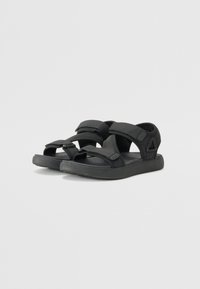 Svarta sandaler i tyg med kardborreband. Har en platt gummisula och en enkel design med öppen tå.