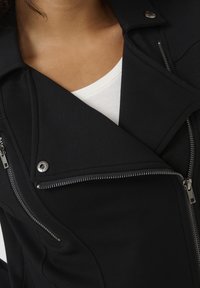 Černá bunda s asymetrickým zipem, zapínáním na patentky, texturovanou tkaninou a stříbrnými kovovými prvky. Má stylový límečkový design.