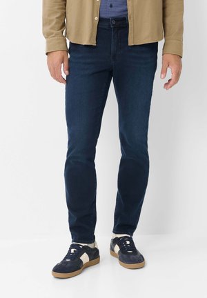 Mann trägt dunkelblaue Slim-Fit-Jeans, marineblaue und weiße Sneaker, beiges Hemd und blau gemustertes Hemd, steht vor weißem Hintergrund.