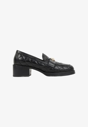 Mocassin en cuir noir matelassé avec talon bloc et emblème décoratif doré à l'avant, présenté de profil sur fond blanc.