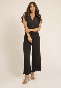 Combinaison noire plissée avec un décolleté en V profond, des manches courtes et des jambes larges. Présente une taille cintrée et un tissu texturé. Accentuée par des talons dorés.