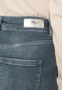 Jeans de denim en un lavado oscuro que presentan una etiqueta plateada con "ONLY" y detalles de costura. La tela parece suave con una ligera textura.