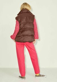 Gilet rempli marron superposé sur un pull en tricot rose, associé à un pantalon rose vif et des baskets grises. Fond minimaliste.