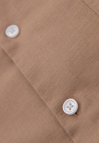 Gros plan d’un tissu beige montrant des boutons blancs à quatre trous cousus sur un tissu texturé avec des coutures et des ourlets visibles.