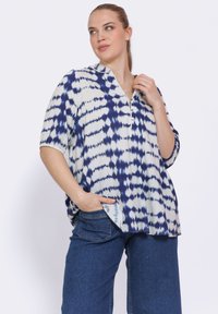 Losse, korteblouse met korte mouwen in het wit met een blauw tie-dye patroon, heeft een V-hals met een gedeeltelijke knoopsluiting en zijzakken.