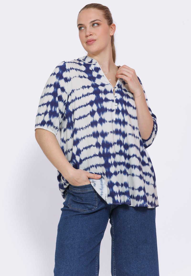 Losse, korteblouse met korte mouwen in het wit met een blauw tie-dye patroon, heeft een V-hals met een gedeeltelijke knoopsluiting en zijzakken.