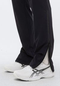 Fekete atlétikai nadrág oldalán cipzárral, fehér tornacipővel párosítva, melyeken fekete részletek és texturált felületek találhatók.