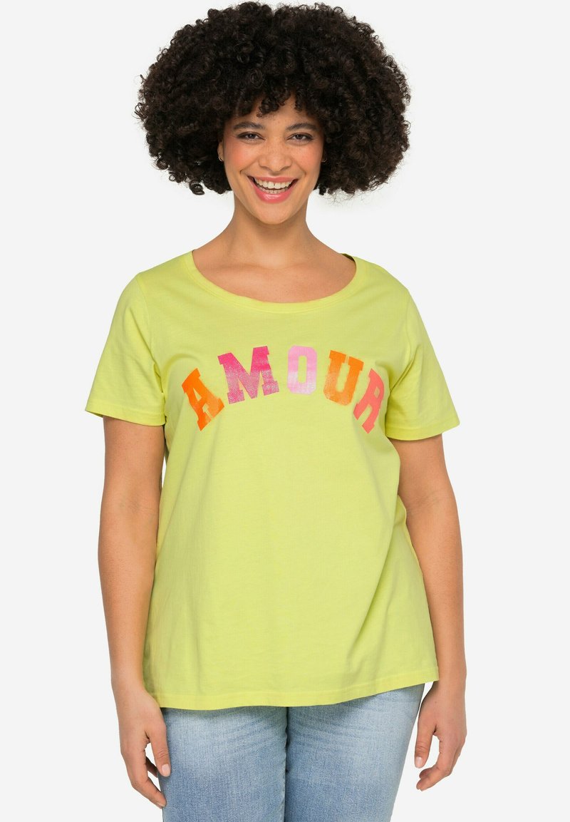 T-shirt di cotone giallo chiaro con scollatura ampia e maniche corte. La scritta "AMOUR" è stampata in lettere vivaci di colore rosa, arancione e rosso.