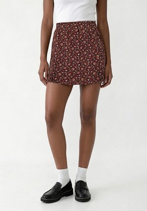 Femme portant une jupe courte marron à fleurs, des chaussettes blanches aux chevilles, des mocassins noirs et un top blanc sans manches, debout devant un fond uni.