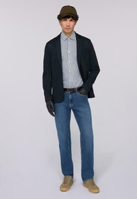 Muž v olivové čepici, černých rukavicích, námořnickém blejzru, pruhované košili s button-up, modrých džínách a béžových teniskách stojí na jednobarevném pozadí.