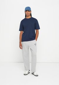 Άτομο που φορά ένα ναυτικό μπλε t-shirt, ανοιχτό γκρι παντελόνι φόρμας με κείμενο, άσπρα και μαύρα αθλητικά παπούτσια και ένα μπλε τζην καπέλο, που στέκεται μπροστά σε λευκό φόντο.