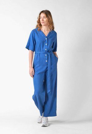 Woman with wavy hair wearing a blue buttoned jumpsuit with a waist tie and white sneakers, standing against a plain white background.