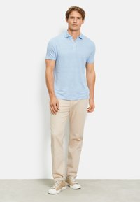 Polo di colore azzurro chiaro con maniche corte e tre bottoni, abbinato a pantaloni color crema e sneakers beige, su uno sfondo neutro.