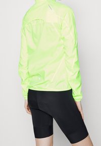 Veste à manches longues légère, réfléchissante de couleur vert néon, avec des poignets élastiques, associée à un cuissard de cyclisme noir ajusté avec texture rembourrée.