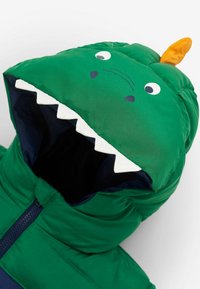 Grön puffjacka med huva designad som en dinosaurie, med vita tänder, blå detaljer och en orange hornapplikation på toppen.