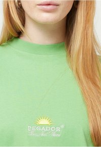 Zielona bawełniana koszulka z ribbowym okrągłym dekoltem. Haftowana z napisem "PEGADOR" oraz grafiką słońca w żółtym i białym kolorze.