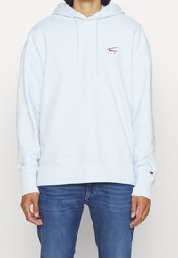 Sudadera con capucha azul claro, con bolsillo canguro frontal, cordones ajustables y un pequeño logo bordado en el pecho. Tela suave.