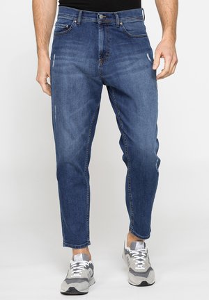 Uomo che indossa jeans blu slim fit con lievi segni di usura e sneakers grigie e bianche, in piedi davanti a uno sfondo semplice.