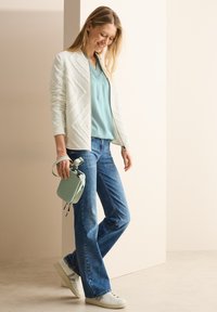 Veste matelassée blanche avec un motif chevron texturé, blouse bleu clair, jean bleu et sac à main rayé vert, accompagnés de baskets blanches.
