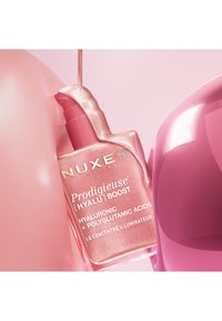 Flacon de sérum NUXE Prodigieuse Hyalu Boost rose avec un liquide scintillant qui s'écoule dessus, sur un fond rose.
