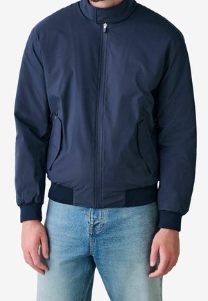 Mann trägt eine marineblaue Bomberjacke mit Reißverschluss, gerippten Ärmelbündchen und Bund, kombiniert mit hellblauen Jeans.