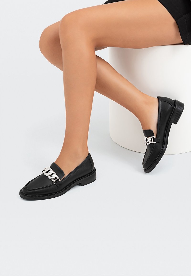 Jambes croisées, portant des mocassins en cuir noir avec un détail de chaîne argentée, assis sur un tabouret cylindrique blanc sur un fond clair.