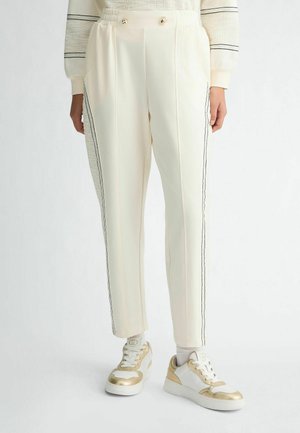 Pantalon beige avec une coupe droite, présentant des rayures noires sur les côtés et deux boutons décoratifs à la taille, associé à des baskets avec des accents dorés.