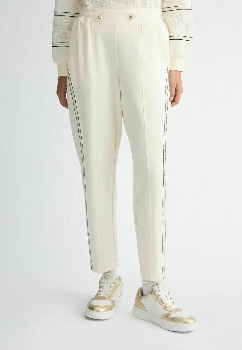 Pantalon beige avec une coupe droite, présentant des rayures noires sur les côtés et deux boutons décoratifs à la taille, associé à des baskets avec des accents dorés.