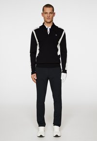 Svart kvartdraget tröja med vita accenter, kombinerad med svarta byxor. Modellen bär vita sportskor och en golfhandske. Slät textur.